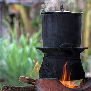 Black pot boiling for the rice cooker on the fired stove next to firewood pile.