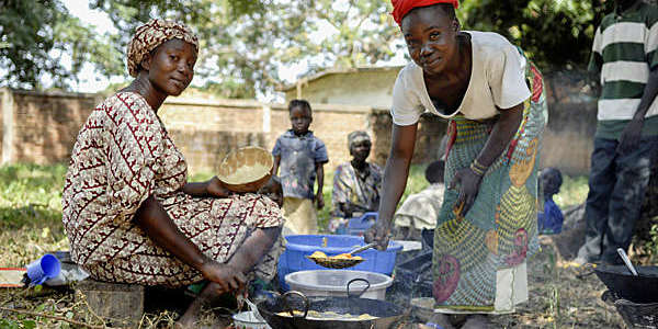 Two African women wearing traditional clothes and colorful headscarves prepare food on an outdoor open fire in Moundou, Chad.