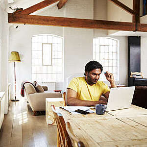 A guy wearing a yellow t-shirt works from home using his dining room table as a desk