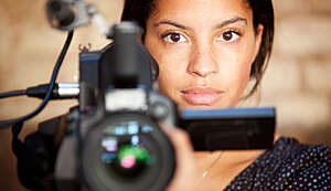 Frontal shot of female video camera operator pointing a camera at her subject, in this case the person viewing the photo.