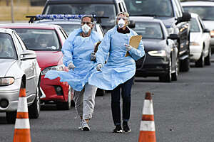 Two female doctors with masks and clipboards walk briskly down a busy road alongside a queue of cars.