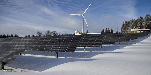Solar panels and wind turbine in a snowy landscape against a bright blue winter sky.